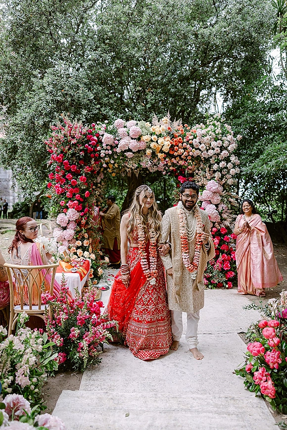 Ceremony recessional as bride and groom walking in red lehenga and sherwani beneath a floral arch, petals on a stone aisle in a garden
