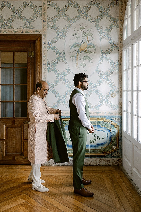 Groom getting ready in an emerald green three piece suit, adjusting his jacket and glasses by tall windows in natural light, patterned wallpaper behind
