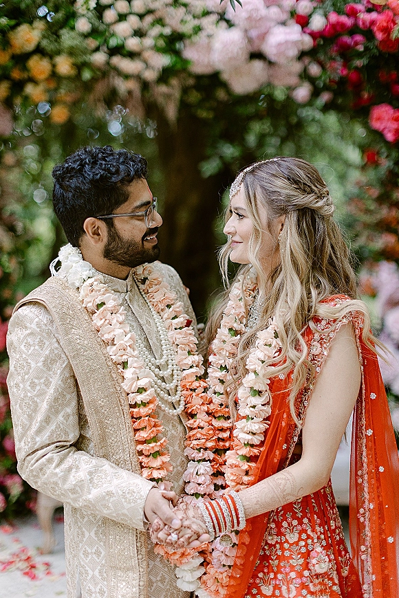 Couple portrait of an Indian wedding couple holding hands and gazing at each other, wearing floral garlands under a greenery arch backdrop