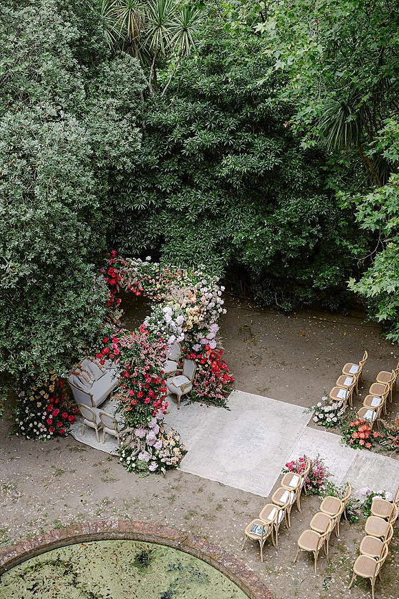 Ceremony setup for an outdoor wedding ceremony with a white aisle runner, floral arch, and lounge seating beside a stone fountain in greenery