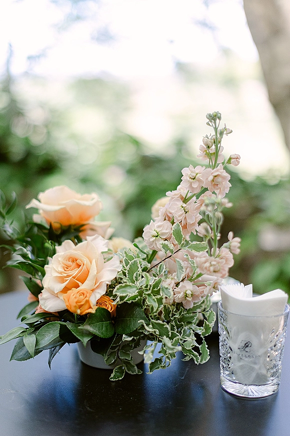 Wedding floral centerpiece with peach roses, blush flowers, and greenery beside a glass tumbler of cocktail napkins in a bokeh garden setting