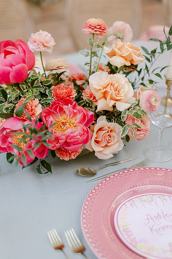 Wedding centerpiece with pink peonies and blush roses, ranunculus and greenery on a white tablecloth with gold flatware and place card