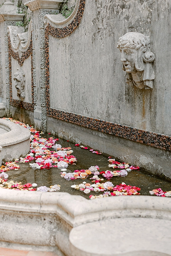 Floating flower petals and floating rose petals drift across a stone fountain basin, creating a romantic water feature centerpiece with blooms