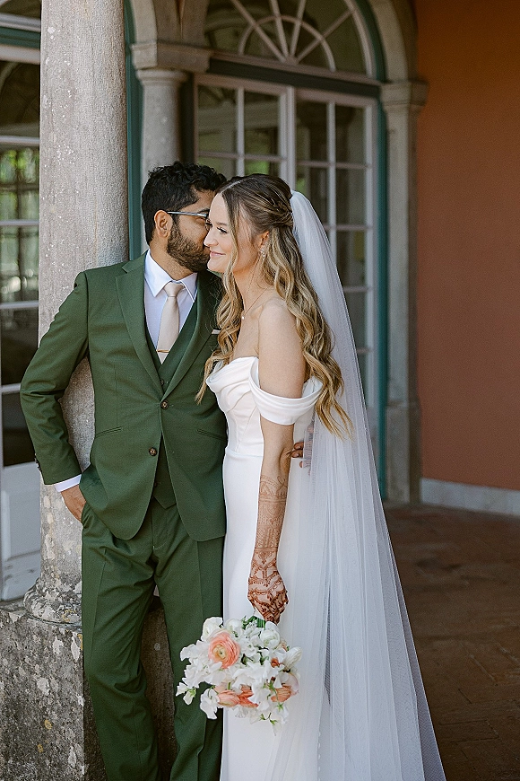 Couple portrait of groom kissing bride’s cheek as she holds a white and peach bouquet, long veil flowing by stone columns and arched windows