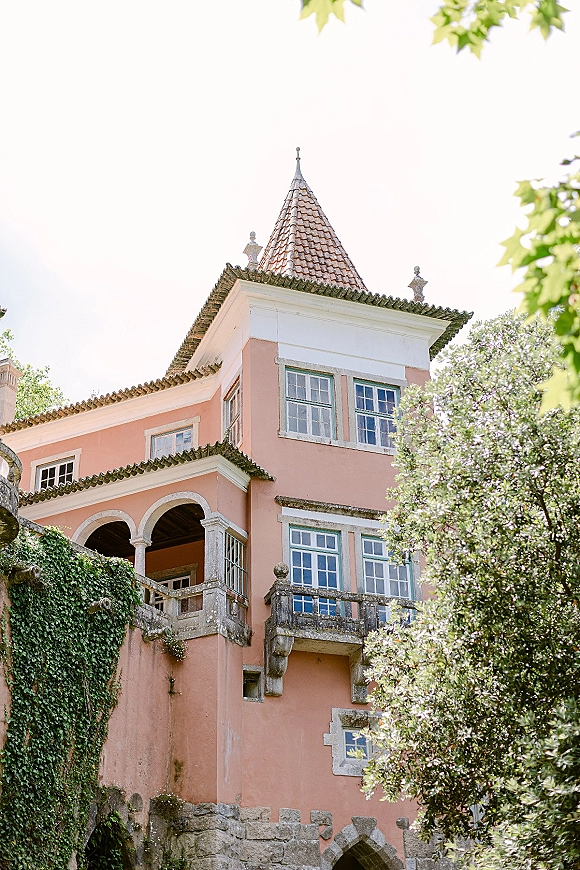Wedding venue exterior of a grand estate wedding venue with tower and arched balcony, tiled roof and ivy vines against trees and sky