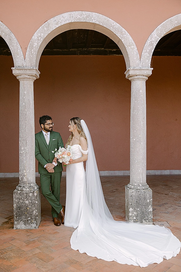 Couple portrait of bride in off shoulder wedding dress with long veil and bouquet facing groom in green suit with glasses under stone arches