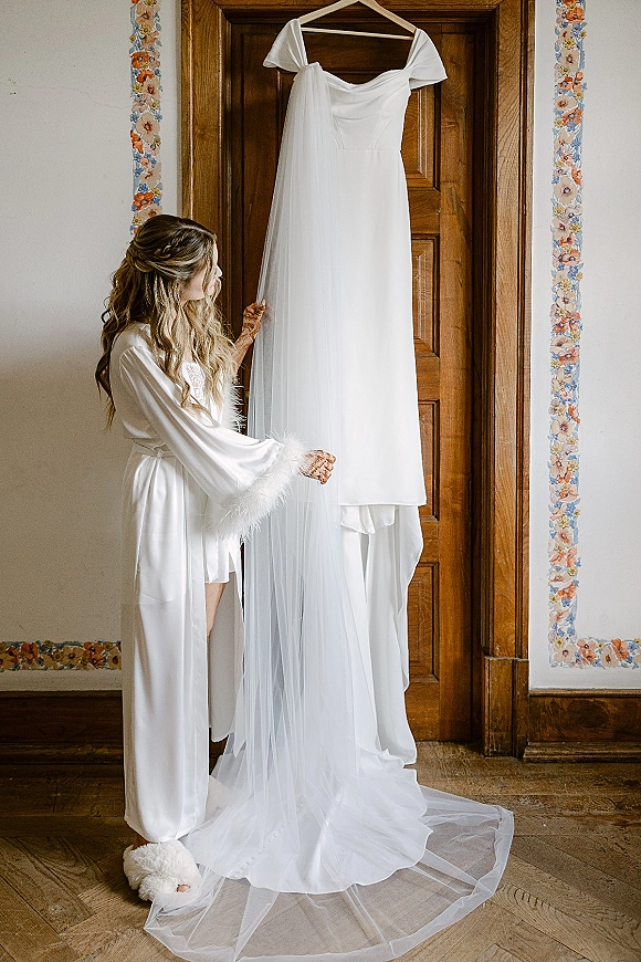 Wedding dress hanging on a hanger with veil draped beside it, framed by a wooden door and floral wallpaper border indoors