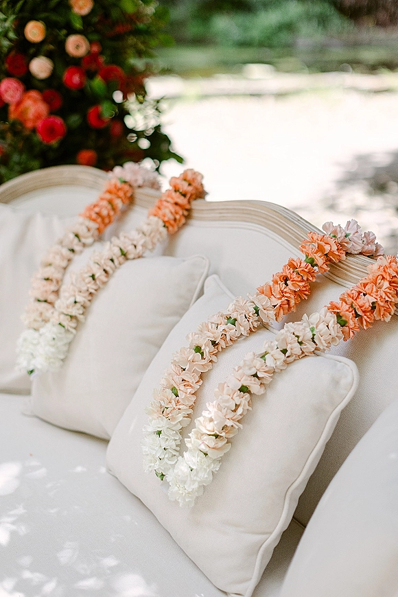 Wedding flower garlands and a floral lei garland draped over a white outdoor sofa with throw pillows in a sunlit garden lounge setting