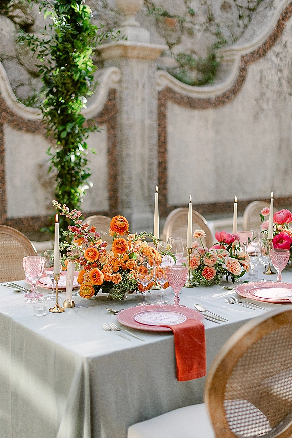 Reception tablescape with wedding table decor featuring orange florals, white taper candles, pink goblets, greenery garland, and a stone wall backdrop