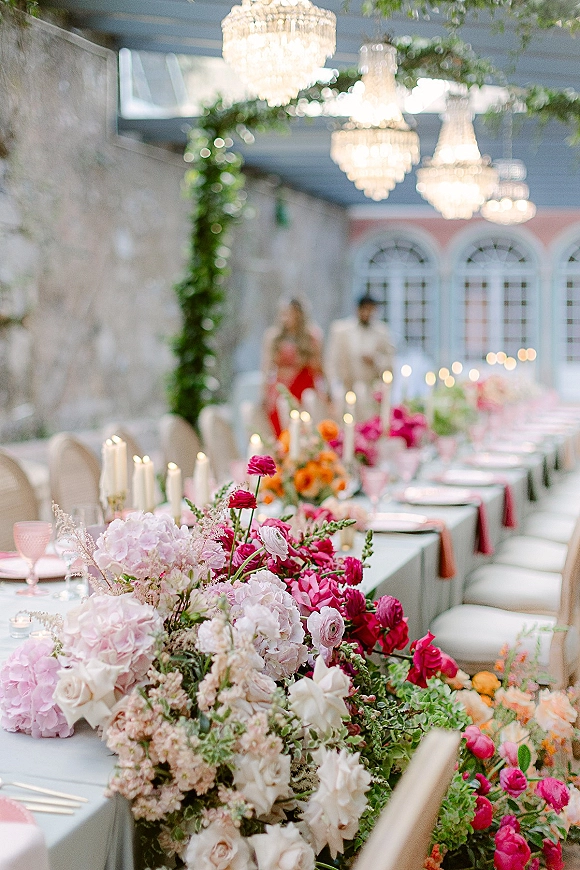 Reception tablescape with long table wedding reception floral runner of pink and orange blooms, taper candles, chandeliers, and arched windows