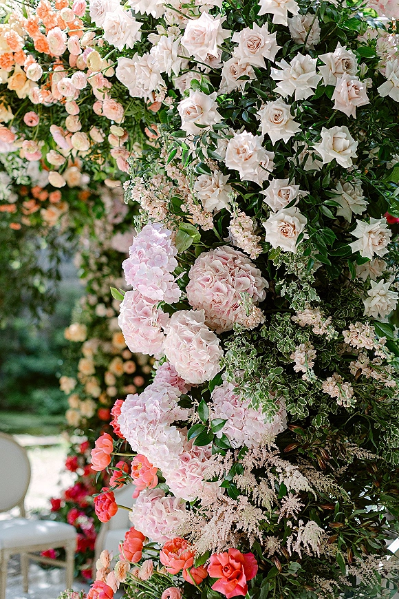 Wedding floral installation framing ceremony chairs, a lush ceremony flower arch of blush roses, hydrangeas, coral blooms, and greenery in a garden setting