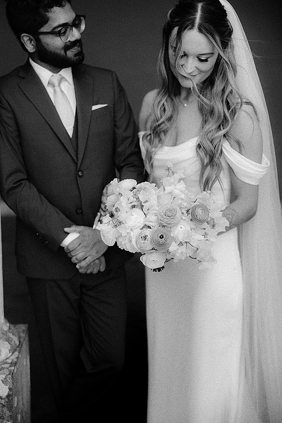 Couple portrait, black and white wedding portrait of groom in glasses looking at bride with veil and white bouquet against a plain wall