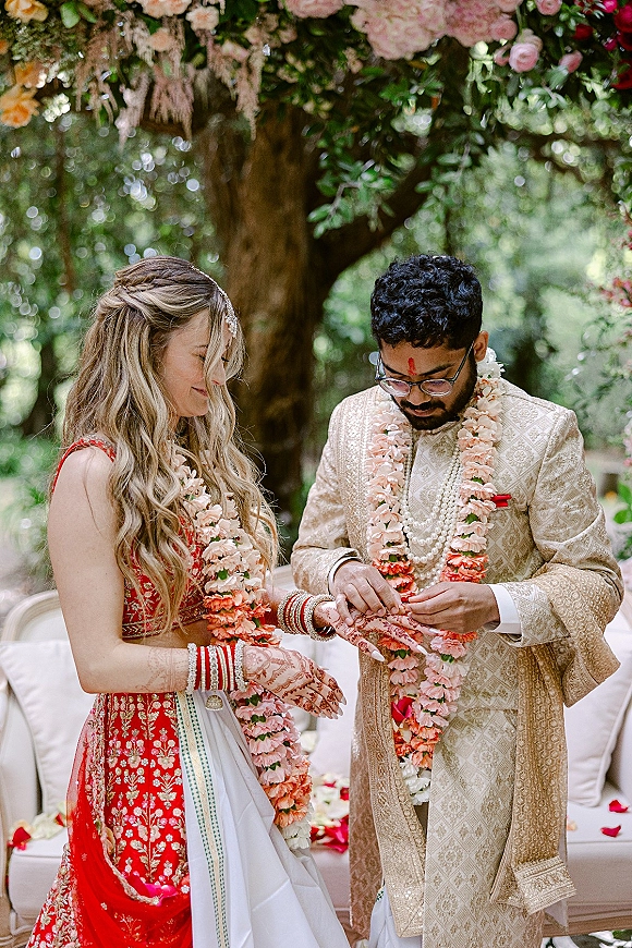 Ceremony moment at an Indian wedding ceremony as bride in red lehenga and groom in sherwani exchange garlands beneath a floral canopy in a garden setting