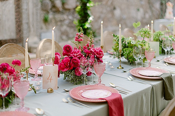 Reception tablescape with wedding table centerpiece of hot pink roses and greenery, taper candles in brass holders on a stone patio table