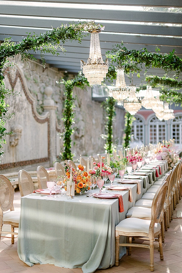 Reception tablescape with long banquet table wedding styling, sage linens, pink goblets, taper candles and florals beneath crystal chandeliers on patio