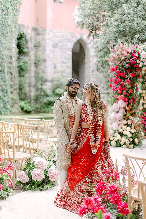 Couple portrait of an Indian wedding couple holding hands under a stone archway in a garden courtyard, bride in a red lehenga with garlands