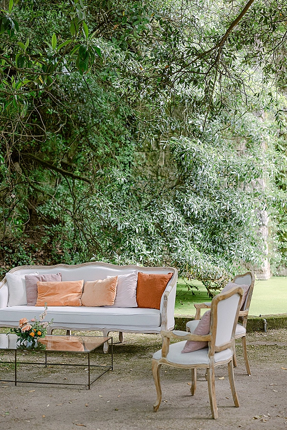 Lounge seating area with vintage sofa and upholstered chairs, throw pillows, and a glass coffee table in a garden setting