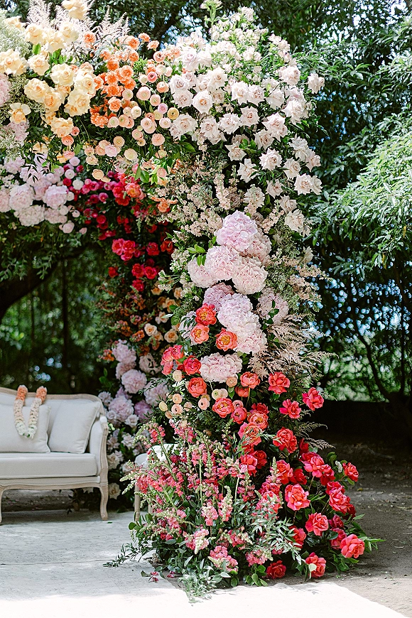Wedding floral arch with roses and peonies, greenery garland, and an upholstered settee with pillows on an outdoor patio by trees