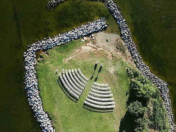 Outdoor ceremony setup with curved rows of white chairs and floral aisle markers on a grassy lawn by a rocky waterfront shoreline