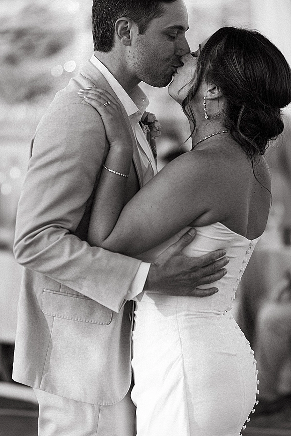 Wedding kiss portrait of bride and groom kiss close up, her strapless gown and jewelry visible as they embrace against outdoor bokeh lights