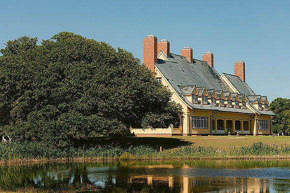 Wedding venue exterior of a yellow manor house with brick chimneys and slate roof, set by a pond with reeds, lawn, and trees under blue sky