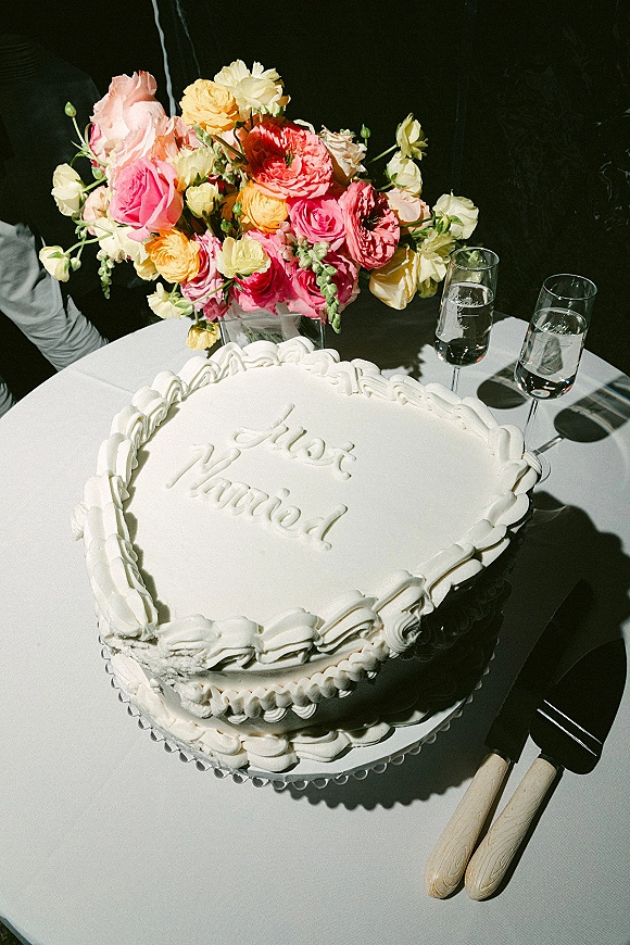 Wedding cake with white buttercream piping and cursive icing writing on a stand beside champagne flutes and roses on a white tablecloth