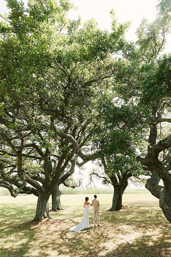 Couple portrait of bride and groom walking away in a strapless dress with long train and light suit beneath sunlit oak trees in a field