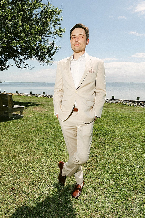 Groom portrait in a beige groom suit with hands in pockets, standing on a grassy waterfront lawn by a tree under blue sky