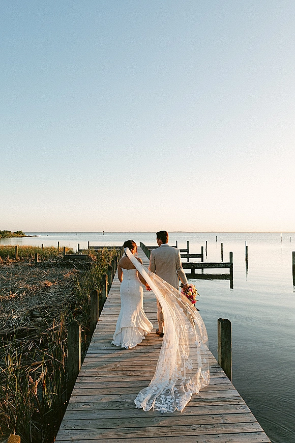 Couple portrait of newlyweds holding hands on a wood dock, bride’s long lace veil blowing behind her over calm marsh water