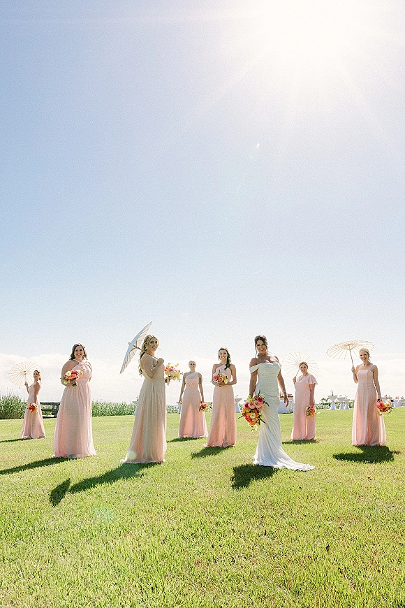 Bridesmaid group photo with bride and bridesmaids in pink dresses holding bouquets and one white parasol on a lawn by the ocean horizon