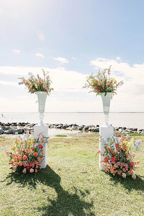 Ceremony floral pillars with wedding floral pillars in tall white urn vases and ground blooms on a grassy waterfront lawn under blue sky