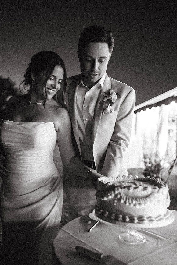 Wedding cake cutting as bride in a strapless gown and groom in a light suit hold a knife at a draped, greenery reception backdrop