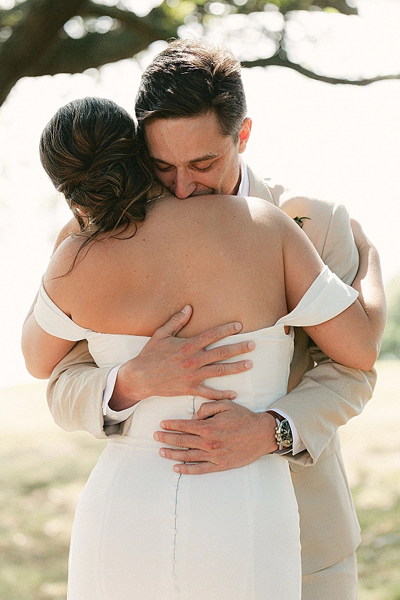 Wedding couple hug as the groom holds the bride from behind, her off-the-shoulder dress visible beneath sunlit trees and bright sky