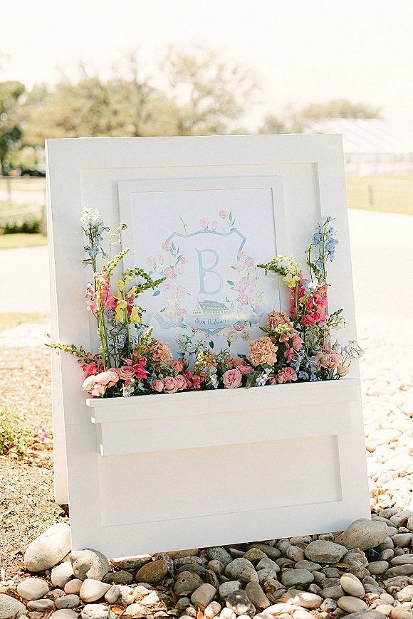 Wedding welcome sign with pastel rose florals at the base, displayed in a white frame on an outdoor lawn with trees and rocks