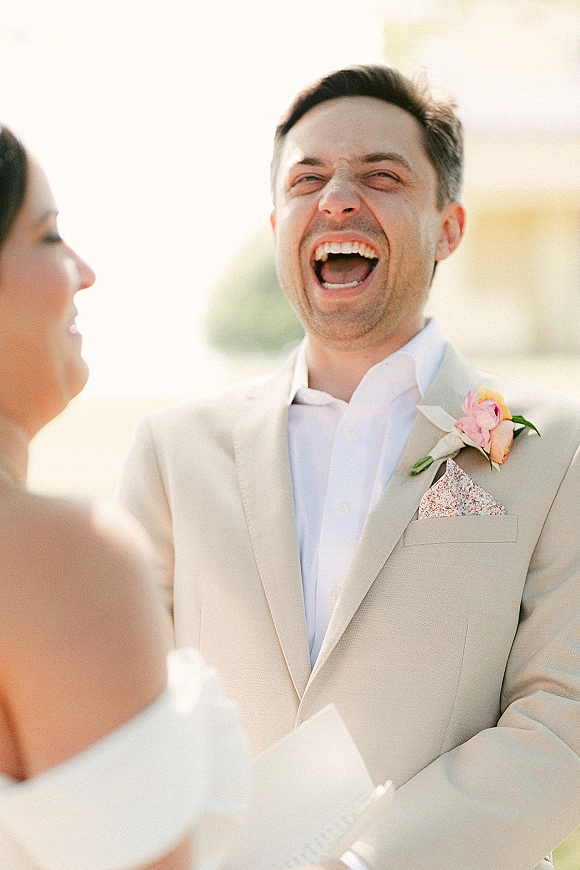 Groom portrait of a groom laughing in a beige suit with pink rose boutonniere and pocket square against bright sky and greenery outdoors