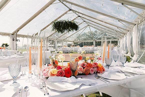 Reception tablescape in a clear tent reception with low floral centerpiece, orange taper candles, blue goblets, and string lights above on the lawn