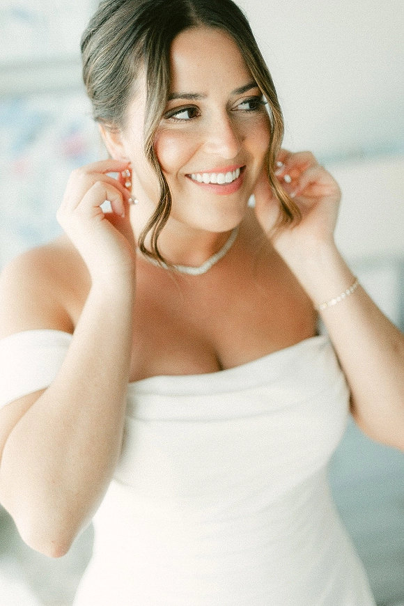 Bridal portrait of a bride putting on pearl earrings in a strapless wedding dress, wearing a delicate necklace against a neutral wall