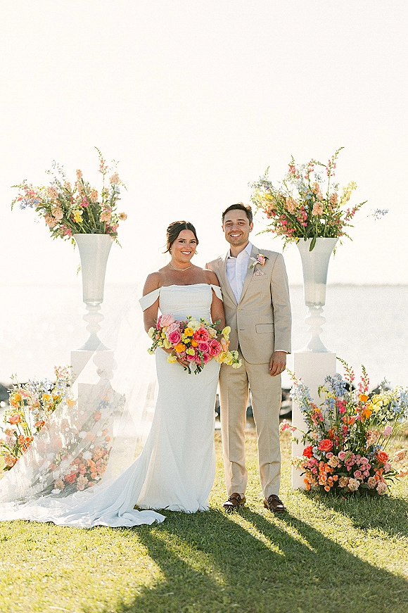 Couple portrait of bride and groom smiling, bride holding colorful bouquet with veil on an oceanfront lawn under bright sky