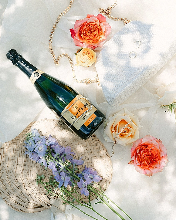Wedding flat lay featuring a champagne bottle, engagement rings on a white woven clutch, roses and purple flowers on soft tulle fabric