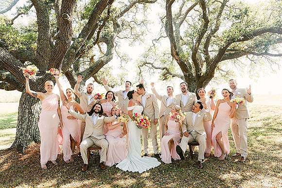 Wedding party portrait with bride and groom kiss as bridesmaids in blush and groomsmen in tan suits cheer under sunlit oak trees