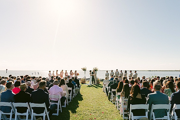 Wedding ceremony on a waterfront lawn with bride and groom at a floral wedding arch, guests in white folding chairs facing the ocean horizon