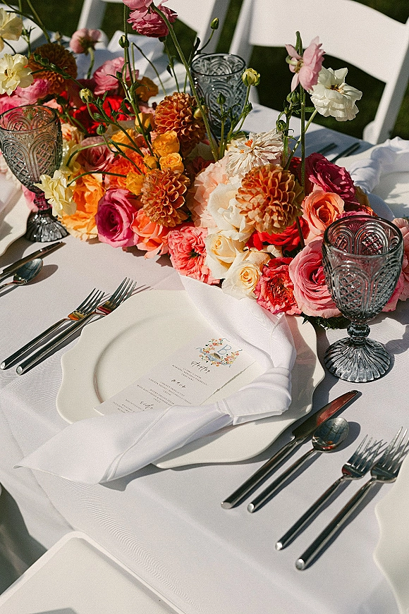 Reception tablescape with wedding table centerpiece of roses and dahlias, menu on plate, blue goblets, silver flatware, and greenery behind white chairs