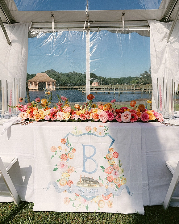 Sweetheart table decor with a sweetheart table floral runner, taper candles, and monogram linen under a clear tent by the waterfront dock