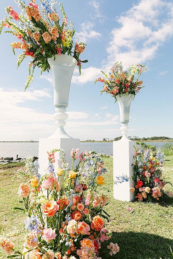 Ceremony floral decor with wedding urn arrangements of roses, delphinium, and greenery on white plinths by a waterfront lawn under blue sky