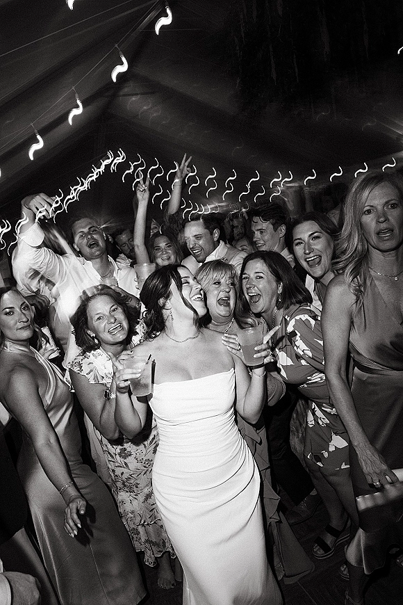 Wedding dance floor packed with bride dancing with guests in a strapless dress, hands up with drinks under string lights in a tent