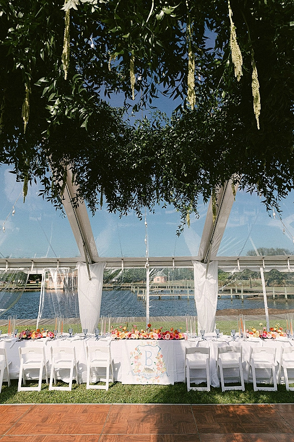 Reception head table with wedding head table decor under a clear-top tent, white draping, greenery, string lights, and waterfront views