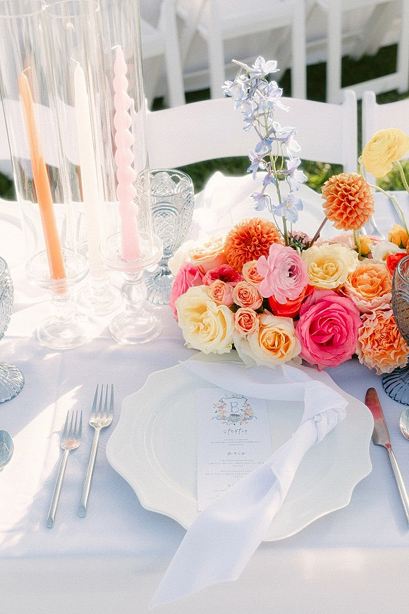 Reception tablescape with colorful wedding tablescape flowers, taper candles, crystal goblets, and scalloped charger plates on a lawn with white chairs