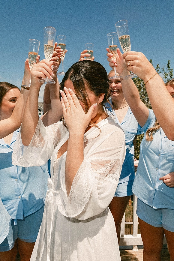 Bridal party toast with bridesmaids champagne toast as bride in lace robe and blue pajamas raise champagne flutes on a sunny deck