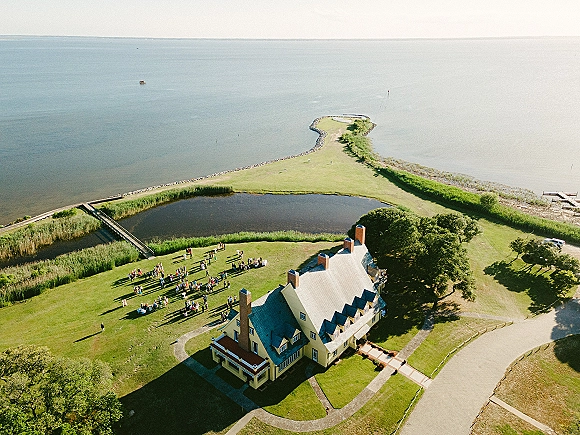 Outdoor wedding ceremony with aerial wedding ceremony view showing guests seated in rows of chairs on a lakeside lawn beside a large house