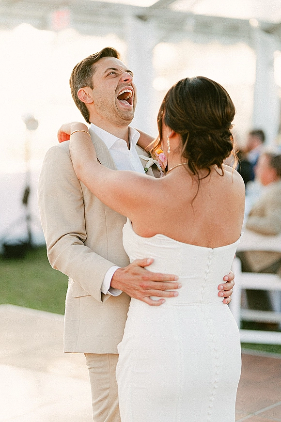 First dance on a wedding dance floor as bride in a strapless gown and groom in a beige suit laugh under string lights in a tent